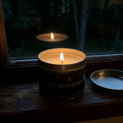 A lit candle in a dark metal tin sits on a wooden windowsill, with the lid placed beside it and the window reflection showing the candle flame. The label reads 'ATTIC WHISpers'.