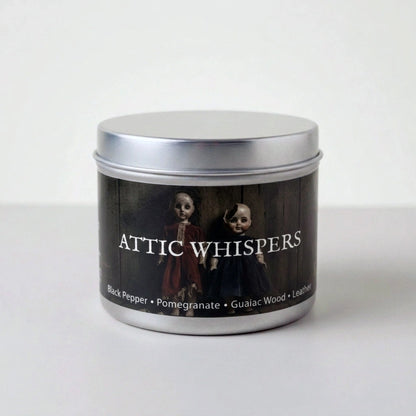 A silver metal tin candle with a black label reading 'ATTIC WHISPERS' and showing two dark-toned figures, photographed on a plain light background.