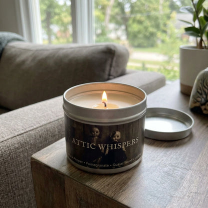 A lit metal tin candle labeled 'ATTIC WHISPER' sits on a wooden table in a cozy living room setting, with its lid placed beside it. A sofa, window, and potted plant are visible in the background.