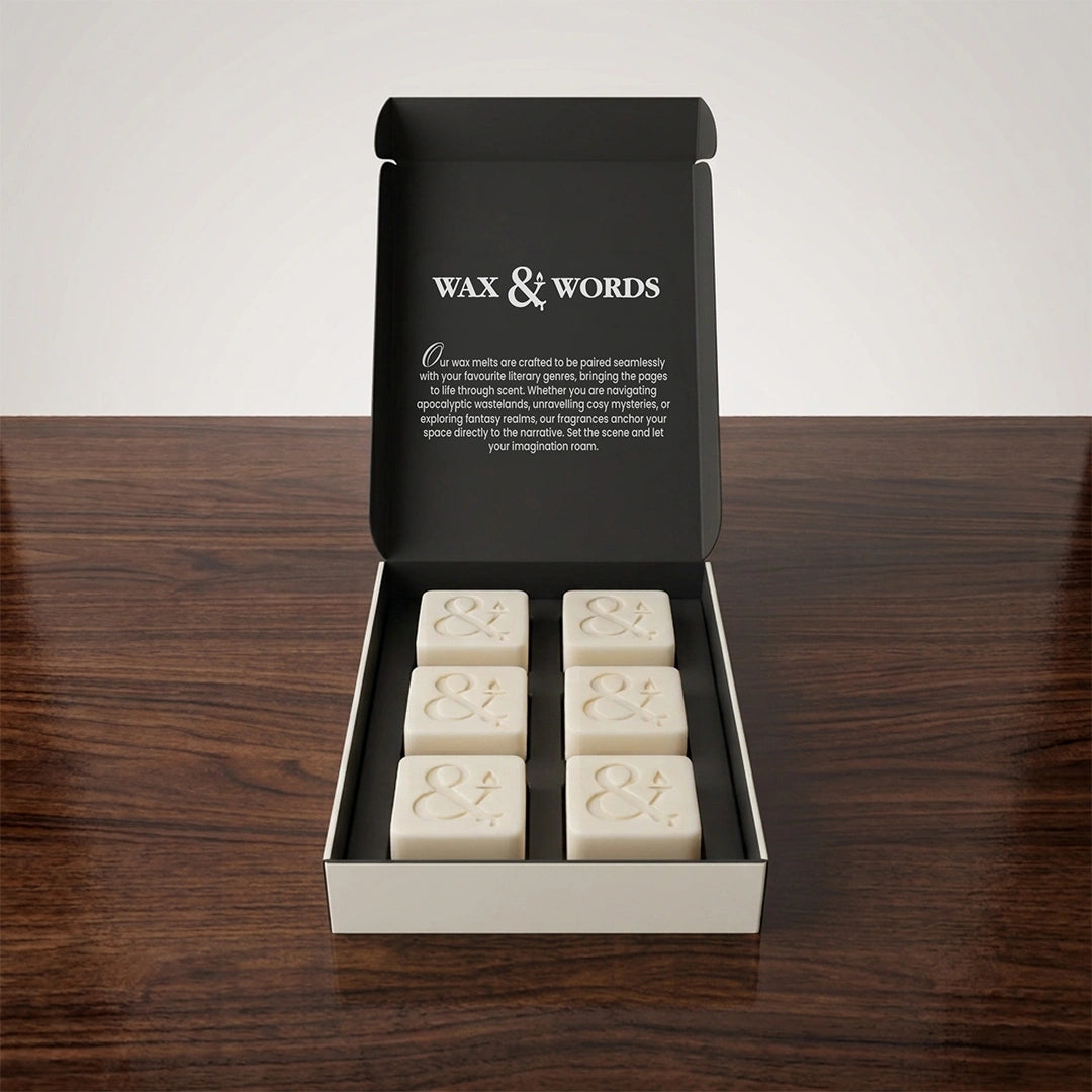 An open black gift box on a dark wooden surface, containing six cream-colored wax melts with embossed ampersands. The inside lid reads 'WAX & WORDS' with descriptive text.