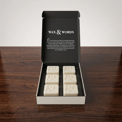 An open black gift box on a dark wooden surface, containing six cream-colored wax melts with embossed ampersands. The inside lid reads 'WAX & WORDS' with descriptive text.