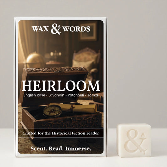 A boxed fragrance product labeled WAX & WORDS and HEIRLOOM stands upright on a light gray surface, with a small off-white wax cube beside it. The box art shows a dark wooden box and compass in a warm interior scene, and the cube has an embossed ampersand symbol.