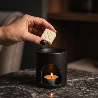 A hand with dark red nail polish holds a small cream-colored wax melt above a black ceramic candle holder with a lit tea light candle inside, set on a dark marble surface.