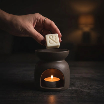 A hand holds a small white rectangular bar with an embossed ampersand above a dark ceramic candle warmer containing a lit tealight candle, set against a dimly lit background.