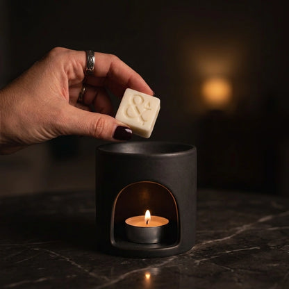 A hand with dark nail polish and a silver ring holds a small cream-colored square soap bar above a black ceramic candle warmer with a lit tea light candle inside, set on a dark marble surface.