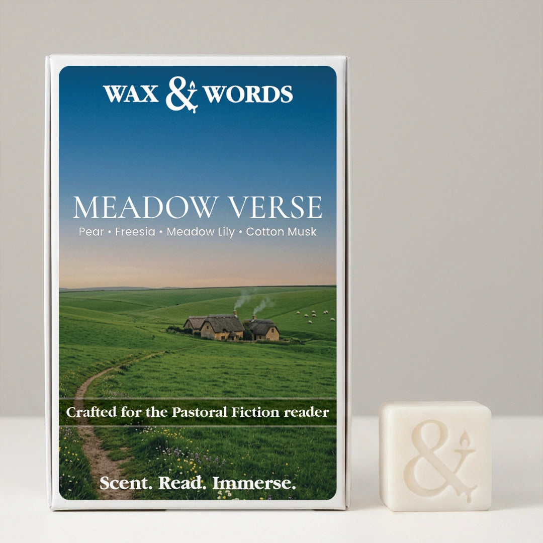 Wax & Words 'Meadow Verse' fragrance box shown upright next to a small off-white bar of the same fragrance on a light gray surface. The box features a blue sky and green field landscape with small houses, and the scent notes Pear, Freesia, Meadow Lily, and Cotton Musk are printed on the front.