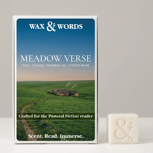 Wax & Words 'Meadow Verse' fragrance box shown upright next to a small off-white bar of the same fragrance on a light gray surface. The box features a blue sky and green field landscape with small houses, and the scent notes Pear, Freesia, Meadow Lily, and Cotton Musk are printed on the front.