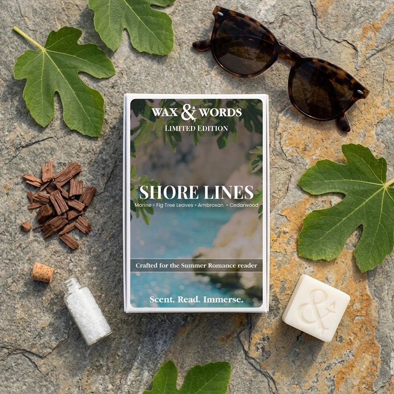 A flat-lay product photograph of the 'Shore Lines' Limited Edition book by Wax & Words. The book is centered on a textured gray stone background, surrounded by thematic props including green fig leaves, a pair of tortoiseshell sunglasses, brown wood chips, a small white bottle, and a white wax melt bar with an ampersand symbol.