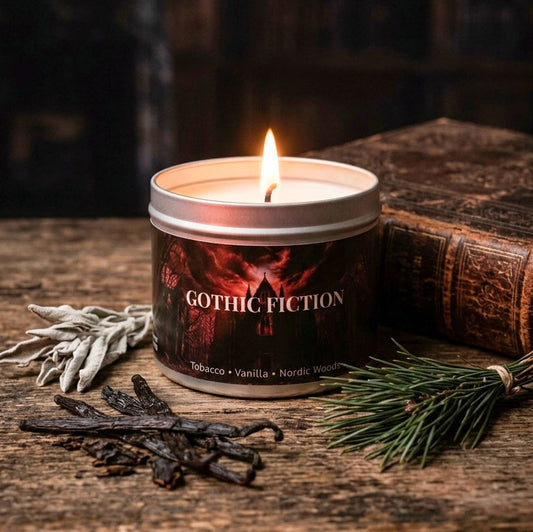 A lit metal tin candle labeled 'Gothic Fiction' sits on a rustic wooden surface with dried herbs, a book, and evergreen branches around it.